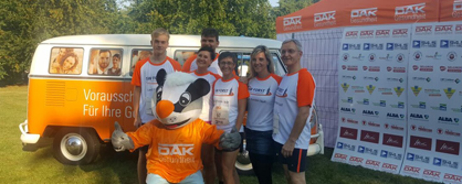 Eine Gruppe Läufer posiert mit dem DAK-Maskottchen vor einem orangenen VW Van