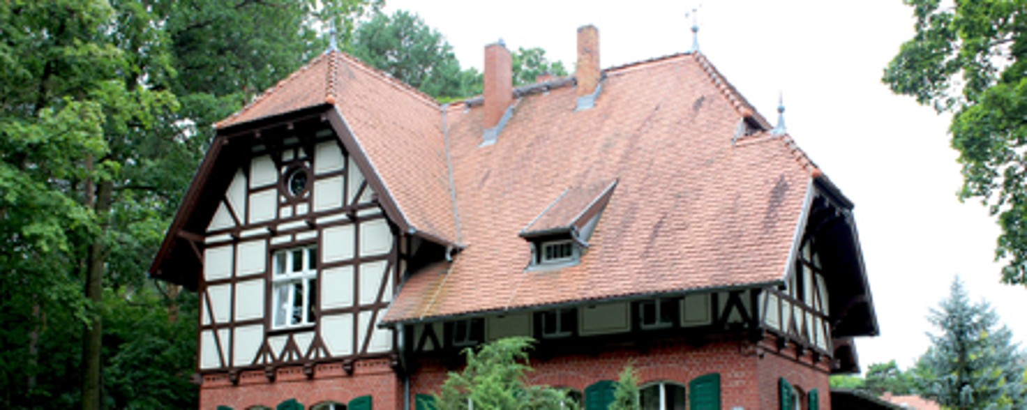 Ein älteres Haus aus Backsteinen und Fachwerkdach steht in einer bewaldeten Umgebung