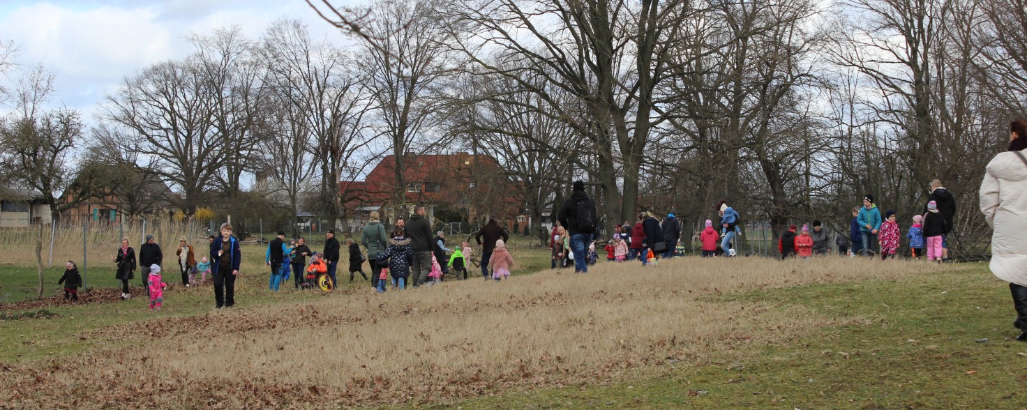 Eine größere Gruppe Menschen mit vielen bunt gekleideten Kindern suchen auf einer Wiese nach Ostereiern