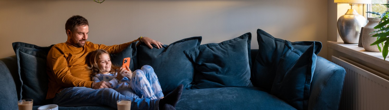 Ein Vater liegt mit seiner kleinen Tochter, die ein Handy in der Hand hält, auf einem gemütlichen Sofa