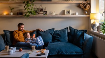 Ein Vater liegt mit seiner kleinen Tochter, die ein Handy in der Hand hält, auf einem gemütlichen Sofa