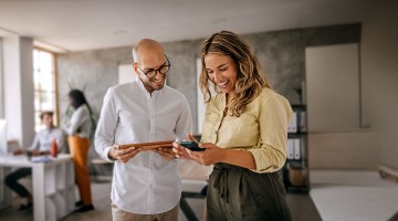 Ein Mann hält ein Tablet in der Hand und schaut lächelnd auf das Handy, das die Frau neben ihm hält